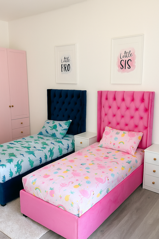 A modern and playful shared kids' bedroom featuring two single beds — one with a deep navy tufted headboard and dinosaur-themed bedding for 'Little Bro', and the other with a bright pink tufted headboard and flamingo-themed bedding for 'Little Sis'. The room includes matching white nightstands with gold knobs and a pastel pink wardrobe, set against a clean white wall with framed prints labeled 'Little BRO' and 'Little SIS' above each bed.