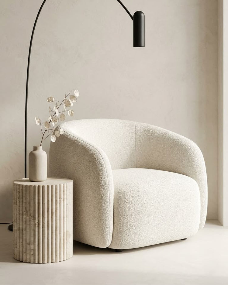 A modern black arched floor lamp stands beside a cream boucle armchair in a minimalist room with a textured beige wall and a small ribbed side table holding a vase with dried branches.